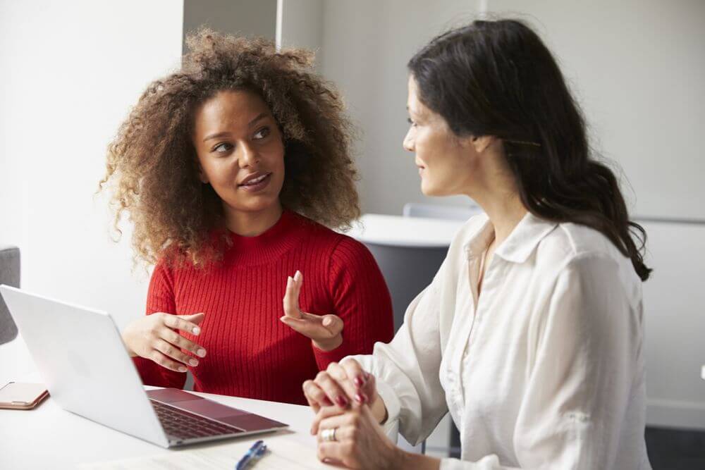 A woman with curly brown hair and a red shirt is speaking with a woman wearing a white shirt who also has brown hair. They are sitting in front of a computer having a meeting