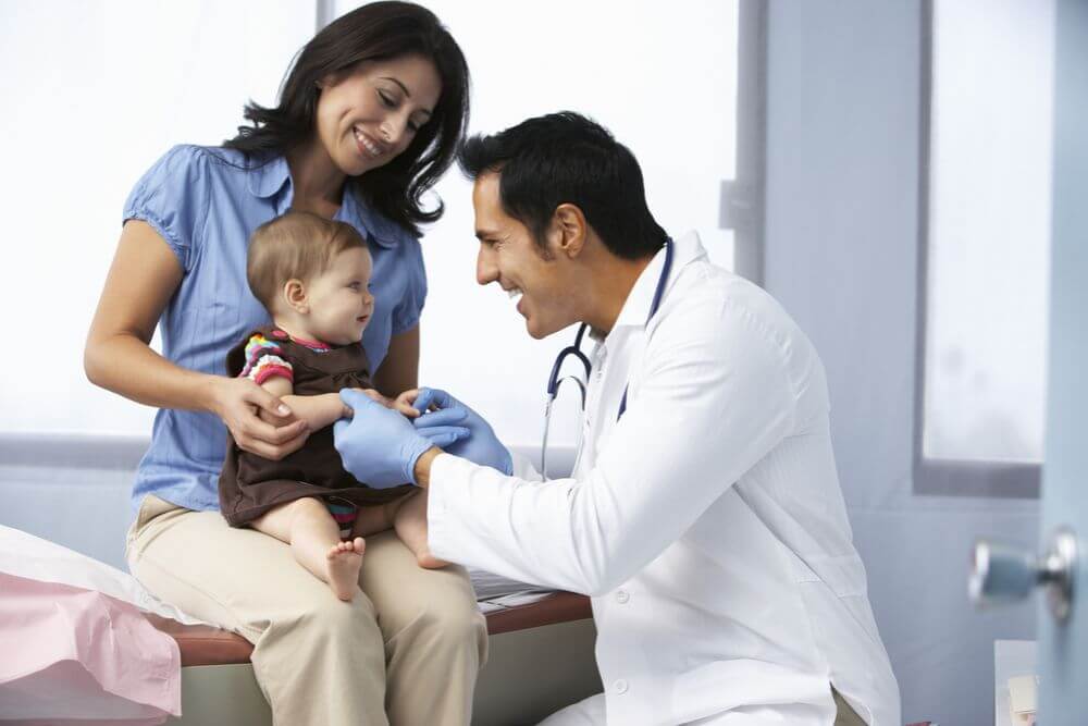 A doctor with a white coat and blue plastic gloves is examining a baby who is being held by a mother wearing a blue shirt and brown pants