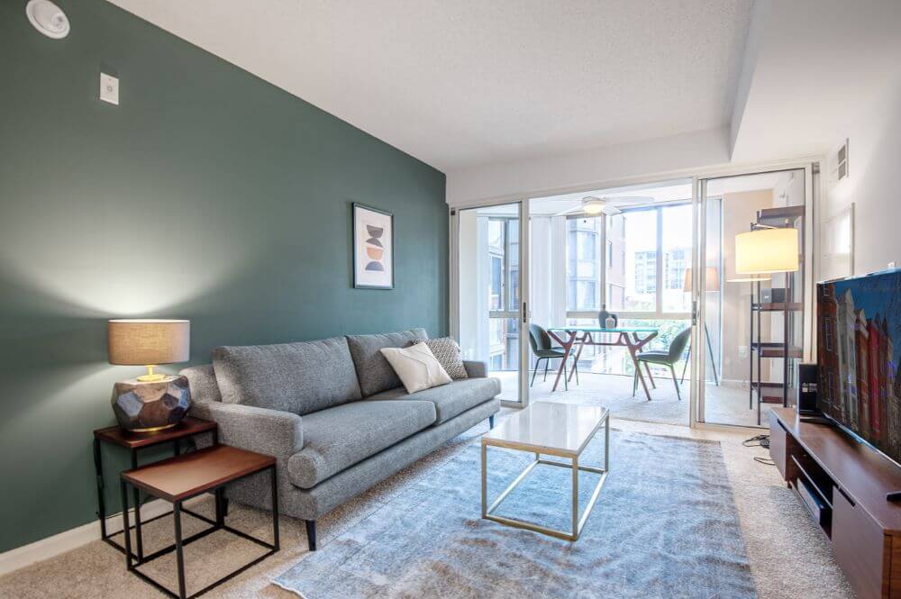 A furnished and equipped apartment in DC that is managed by Blueground. There is a dark green wall behind a grey couch as well as a coffee table, TV and a dining area to the right