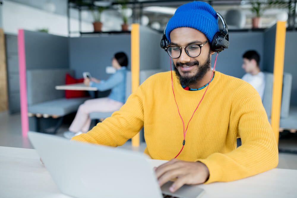 A man wearing a yellow sweater and a blue heat and black headphones is working on a silver laptop while behind him people are sitting in individual meetings room within a coworking space
