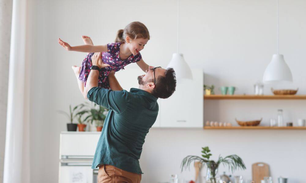 A father wearing a green shirt and brown pants is lifting his daughter in the air and playing with her while they both smile