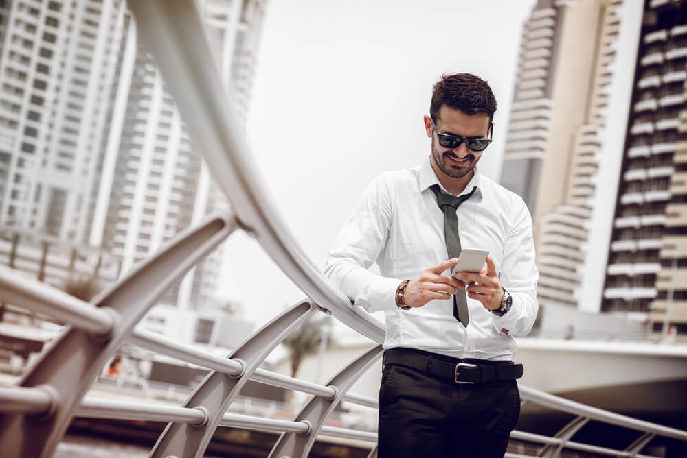 A man wearing formal clothes, looking at his mobile phone while leaning against a railing in Dubai