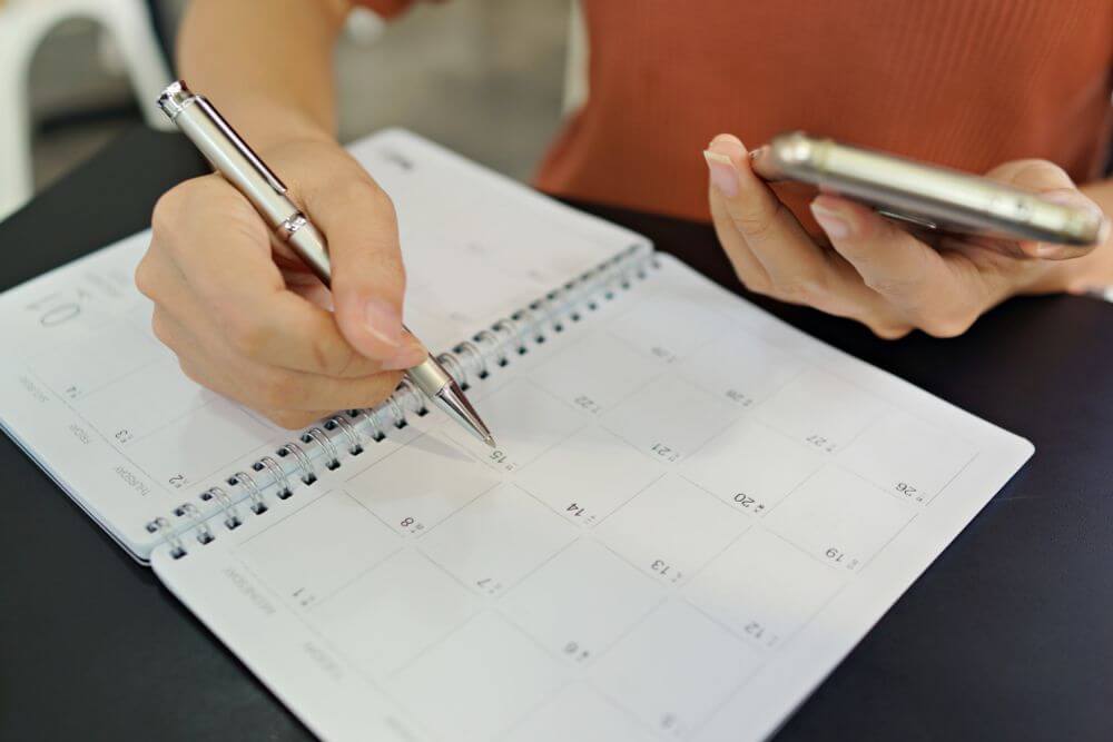 A woman wearing an orange shirt and holding her mobile phone in one hand and a pen in the other. She has a calendar in front of her and is keeping track of a timeline
