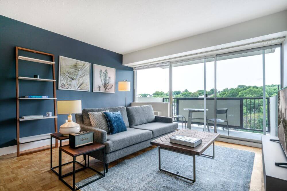 A furnished and equipped apartment in Boston managed by Blueground. There is a dark blue accent wall and a grey couch in the center of the room. There is a balcony to the left of the couch with glass across the entire wall.