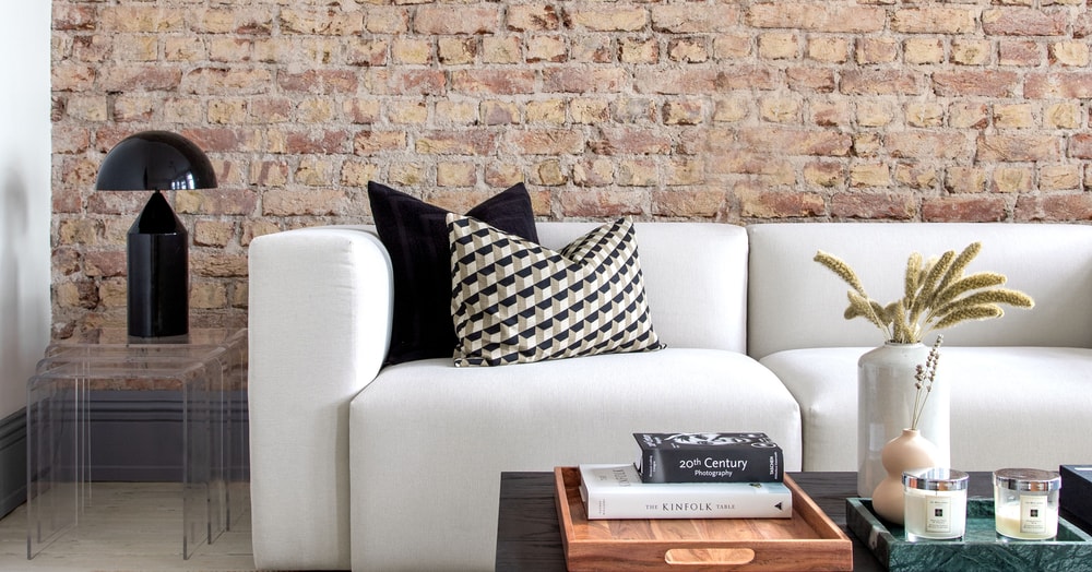 white couch with a black pillow and a black and white pillow against an exposed brick wall with a table in front of it with books and a vase with a black lamp on the side