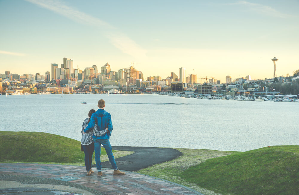 A man and a woman stand together with their arms around each other facing the Seattle sky line