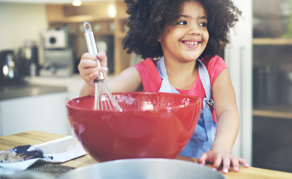 A girl with big black curly hair is holding a whisk and mixing ingredients together in a bowl in the kitchen