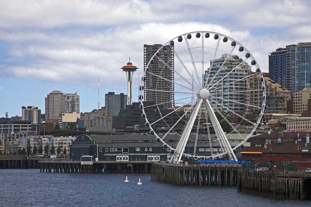 A large white ferris wheel in Seattle near the waterfront. There are old buildings all around and the Space Needle can be seen sticking out against the thick layer of white clouds