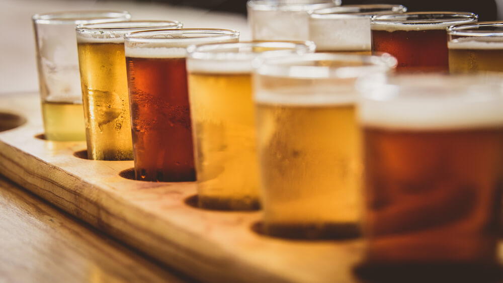 A flight of multicolored beers sits in a wooden holder on a wooden table at a brewery