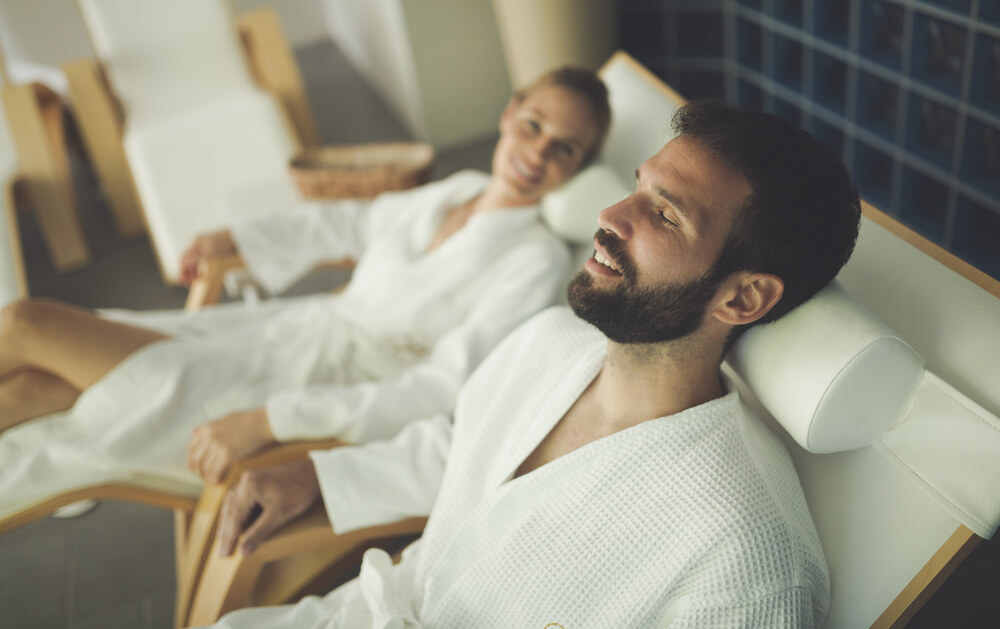 man with dark brown hair an beard wearing a white robe is laying down with his eyes closed feeling relaxed next to a woman who is wearing a white robe and smiling. They are in a spa