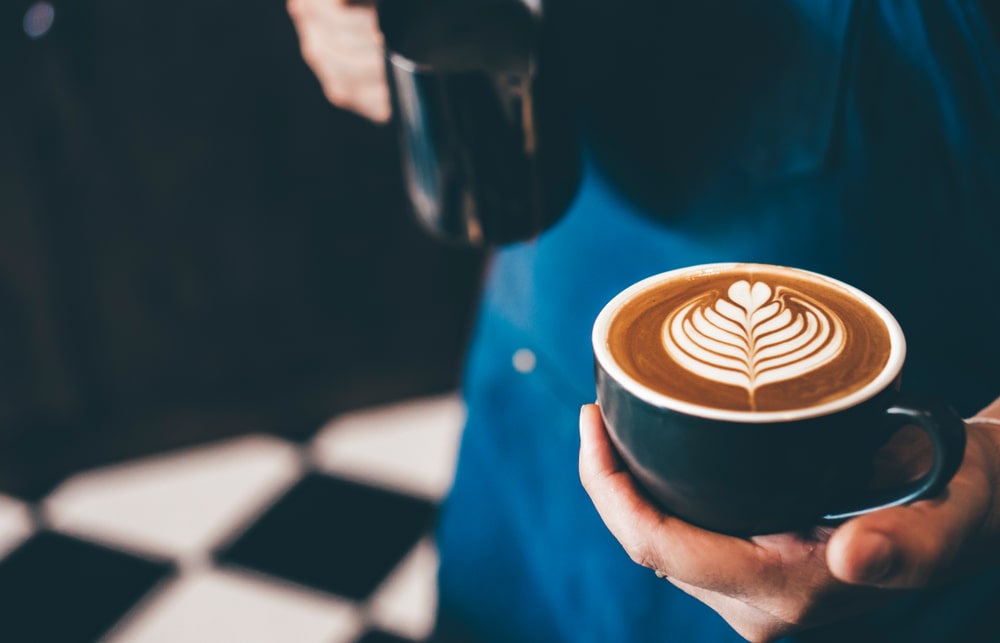 Unknown person with blue apron holding a blue cup of coffee with latte art in their hand with chequered black and white tiles in the background
