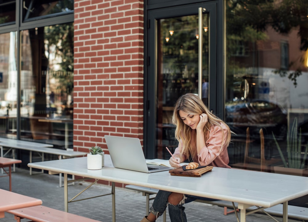 Blonde girl wearing a pink shirt sitting outside on a long white table with her elbow on the table and her hand on her face while writing in her notebook with a laptop and coffee cup next to her
