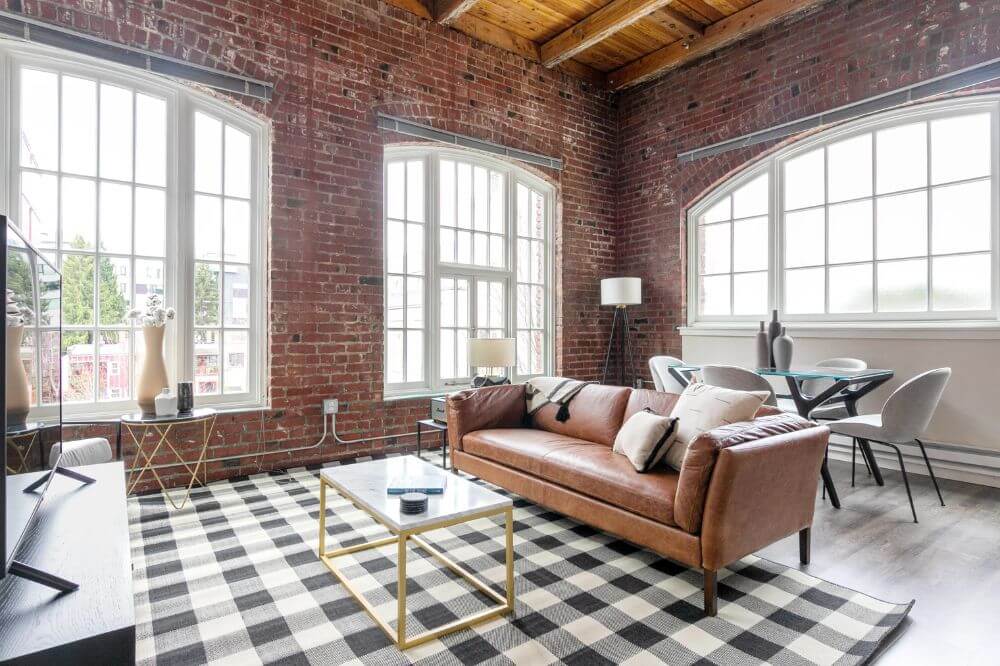 a leather sofa next to a coffee table on top of a black and white chequered carpet inside a brick wall apartment with three big windows with white window panes 