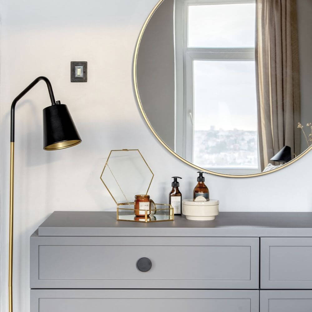 A close up look at a greyish blue dresser with a round mirror above and a standing lamp to the side. On top of the dresser are some small amenities for Blueground guests to enjoy such as high quality creams.