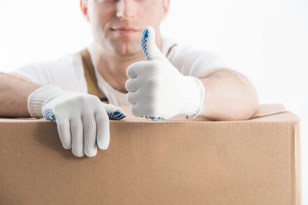 A man holding a cardboard box and wearing white gloves and facing forward giving a thumbs up sign