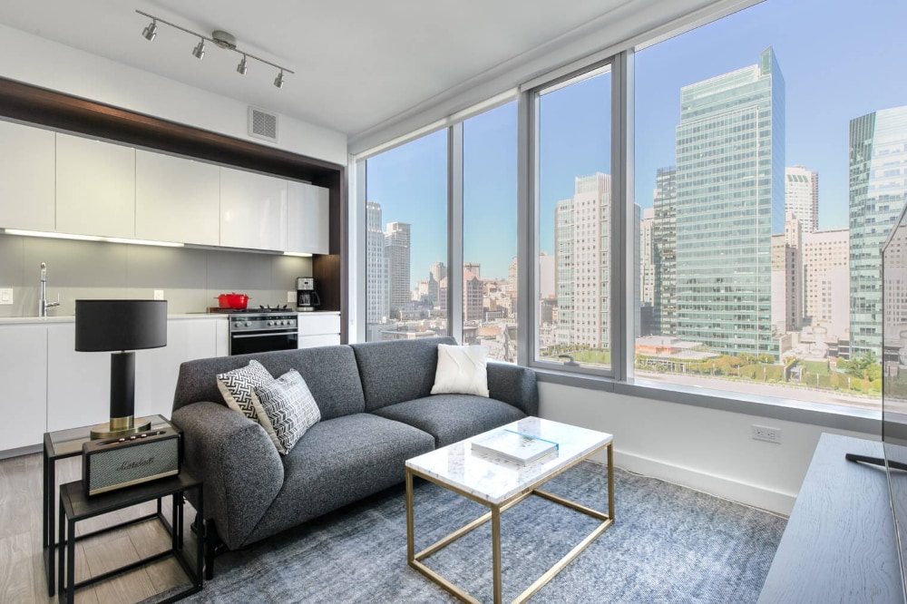 Panoramic view of interior of an apartment with a clear blue sky view of the San Francisco skyline. Inside the apartment there is a grey sofa with a small marble coffee table in front it and a grey carpet underneath. Behind there is a white modern kitchen with kitchen appliances. 