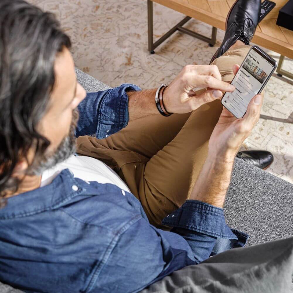 A man with dark black hair wearing a white shirt and a denim jacket and brown pants is sitting on a grey couch in front of a wooden coffee table. He has on black shoes and is wearing a watch. He is in the middle of using the Blueground Guest App on his mobile phone.