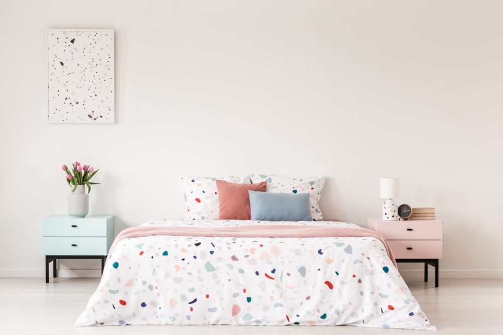 a bedroom with white walls and pastel green night table on the left side of the bed and a pastel pink night table on the right side of the bed. The bed is large with a white cover with a multicolored, pastel modern design.