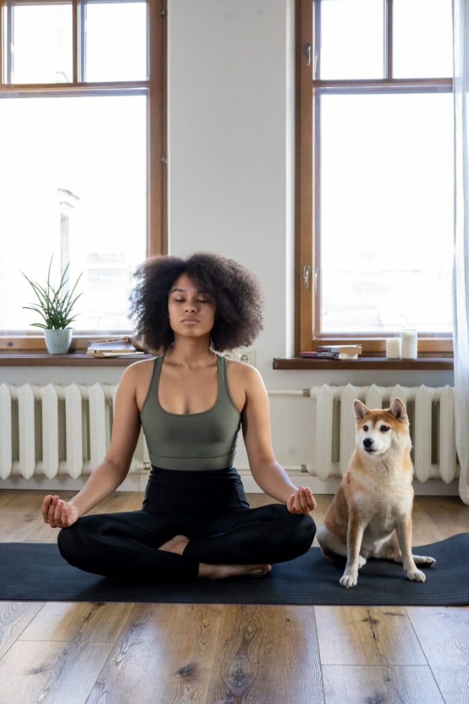 a girl is meditating on a yoga mat while her dog is standing next to her